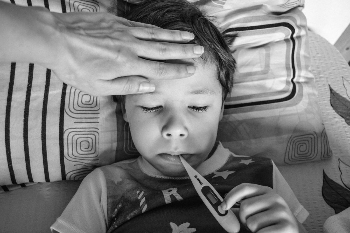 Bé trai nằm nghỉ, tay mẹ đặt lên trán, nhiệt kế ngậm trong miệng